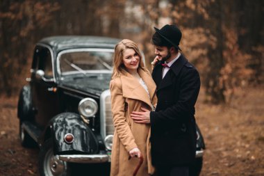 Gorgeous newlywed bride and groom posing in pine forest near retro car in their wedding day