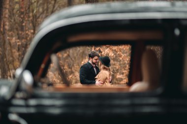 Gorgeous newlywed bride and groom posing in pine forest near retro car in their wedding day