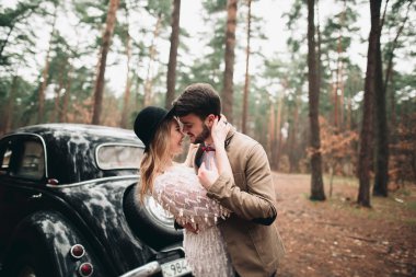 Gorgeous newlywed bride and groom posing in pine forest near retro car in their wedding day