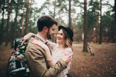Gorgeous newlywed bride and groom posing in pine forest near retro car in their wedding day