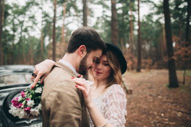 Gorgeous newlywed bride and groom posing in pine forest near retro car in their wedding day