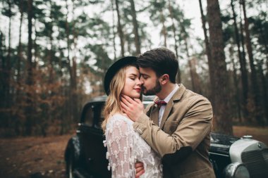 Gorgeous newlywed bride and groom posing in pine forest near retro car in their wedding day