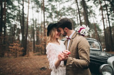 Gorgeous newlywed bride and groom posing in pine forest near retro car in their wedding day