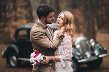 Gorgeous newlywed bride and groom posing in pine forest near retro car in their wedding day