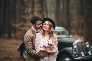 Gorgeous newlywed bride and groom posing in pine forest near retro car in their wedding day