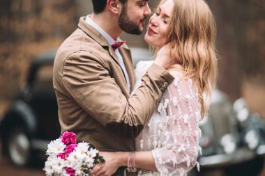 Gorgeous newlywed bride and groom posing in pine forest near retro car in their wedding day