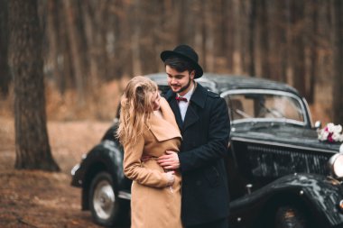 Gorgeous newlywed bride and groom posing in pine forest near retro car in their wedding day