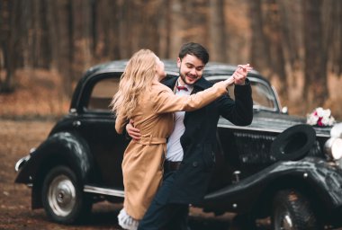 Gorgeous newlywed bride and groom posing in pine forest near retro car in their wedding day