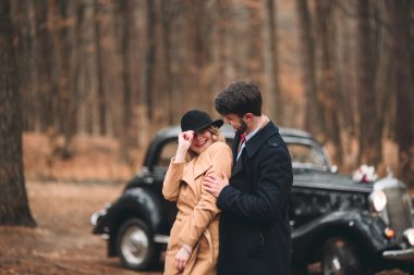 Gorgeous newlywed bride and groom posing in pine forest near retro car in their wedding day