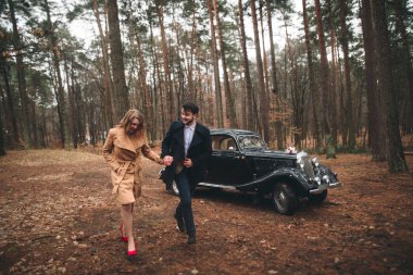 Gorgeous newlywed bride and groom posing in pine forest near retro car in their wedding day