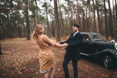Gorgeous newlywed bride and groom posing in pine forest near retro car in their wedding day