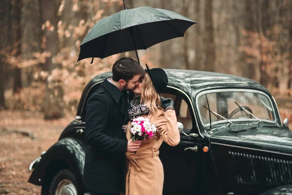 Gorgeous newlywed bride and groom posing in pine forest near retro car in their wedding day