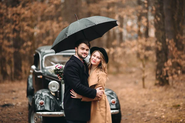 Gorgeous newlywed bride and groom posing in pine forest near retro car in their wedding day — Φωτογραφία Αρχείου