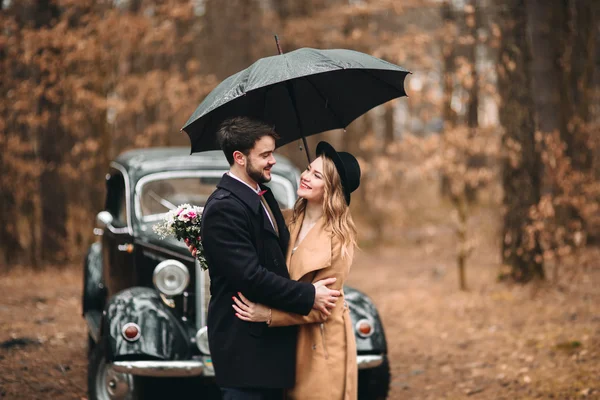 Gorgeous newlywed bride and groom posing in pine forest near retro car in their wedding day — ストック写真
