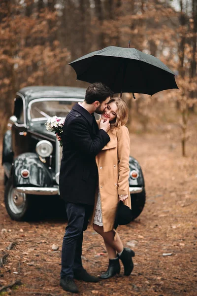 Gorgeous newlywed bride and groom posing in pine forest near retro car in their wedding day