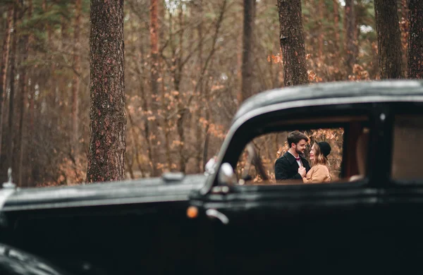Великолепные молодожёны позируют в сосновом лесу возле ретро-машины в день свадьбы — стоковое фото