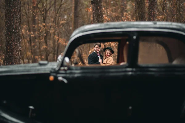 Linda noiva recém-casada e noivo posando na floresta de pinheiros perto do carro retro em seu dia do casamento — Fotografia de Stock