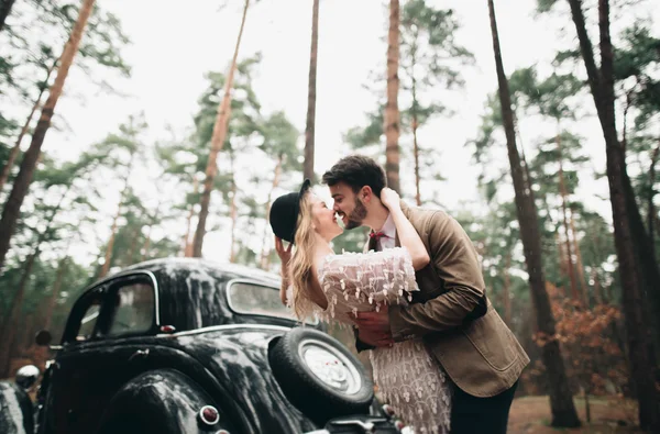 Gorgeous newlywed bride and groom posing in pine forest near retro car in their wedding day — Φωτογραφία Αρχείου
