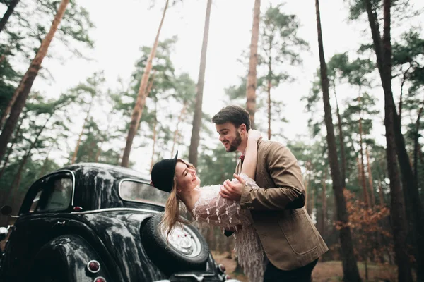 Hermosa novia recién casada y novio posando en un bosque de pinos cerca de coche retro en el día de su boda — Foto de Stock