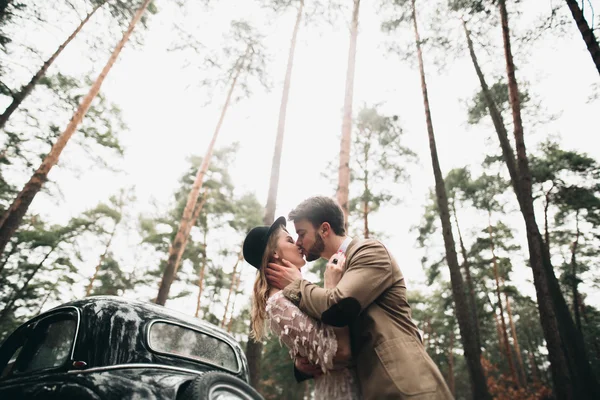 Gorgeous newlywed bride and groom posing in pine forest near retro car in their wedding day — Φωτογραφία Αρχείου