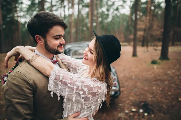 Gorgeous newlywed bride and groom posing in pine forest near retro car in their wedding day — 图库照片