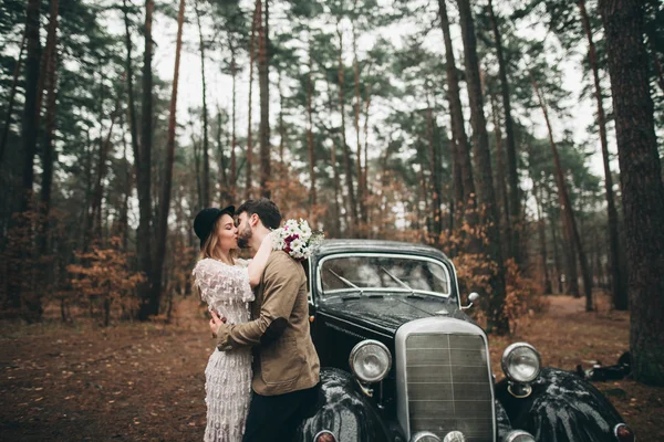 Gorgeous newlywed bride and groom posing in pine forest near retro car in their wedding day — Stok fotoğraf