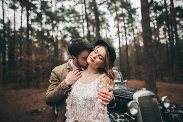 Gorgeous newlywed bride and groom posing in pine forest near retro car in their wedding day — Stock fotografie
