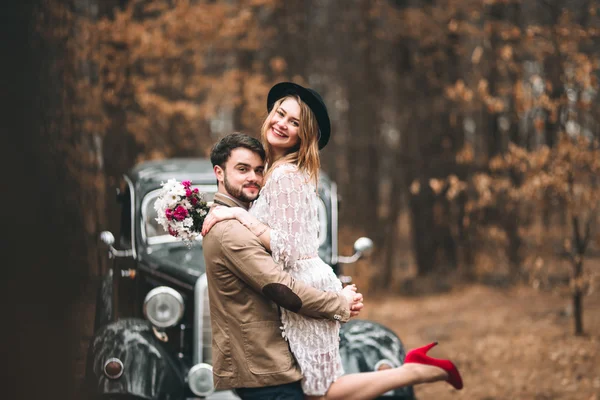 Hermosa novia recién casada y novio posando en un bosque de pinos cerca de coche retro en el día de su boda — Foto de Stock
