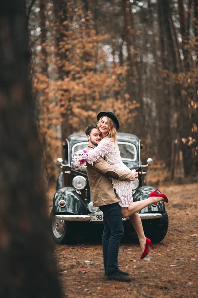 Gorgeous newlywed bride and groom posing in pine forest near retro car in their wedding day