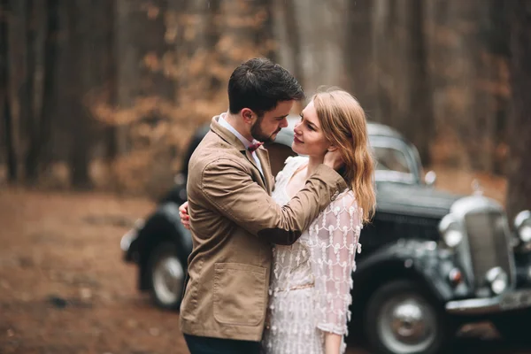 Gorgeous newlywed bride and groom posing in pine forest near retro car in their wedding day — Stock Fotó