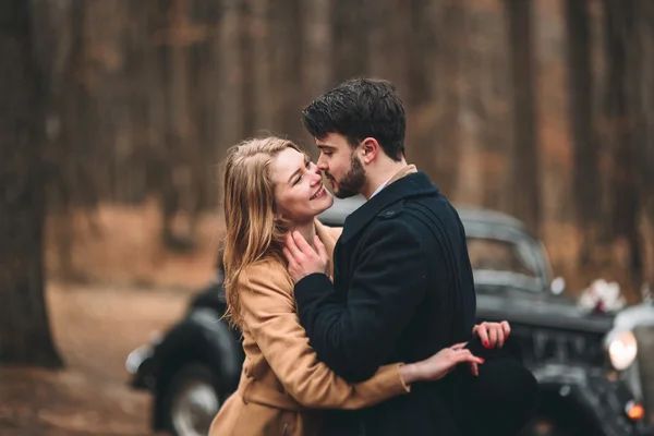 Gorgeous newlywed bride and groom posing in pine forest near retro car in their wedding day — 스톡 사진