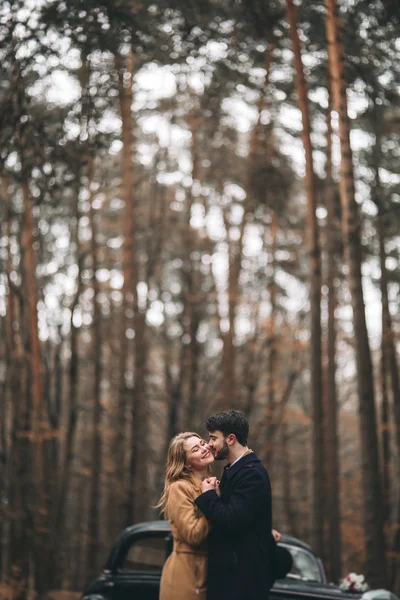 Gorgeous newlywed bride and groom posing in pine forest near retro car in their wedding day — Stockfoto