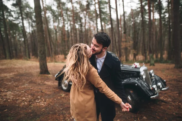 Gorgeous newlywed bride and groom posing in pine forest near retro car in their wedding day — Φωτογραφία Αρχείου