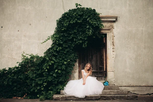 Hermosa joven novia rubia con ramo de novia sentado en las escaleras bajo plantas magníficas — Foto de Stock
