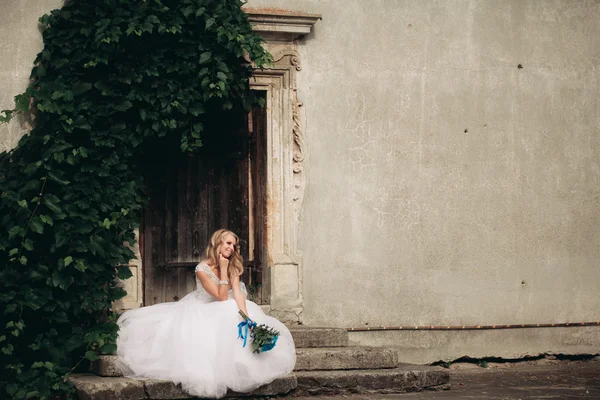 Belle jeune mariée blonde avec bouquet nuptiale assise dans les escaliers sous de magnifiques plantes — Photo