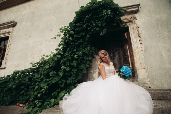 Mooie jonge blonde bruid met bruids boeket zittend op de trap onder prachtige planten — Stockfoto