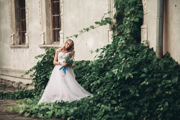 Hermosa joven novia rubia con ramo de novia sentado en las escaleras bajo plantas magníficas — Foto de Stock