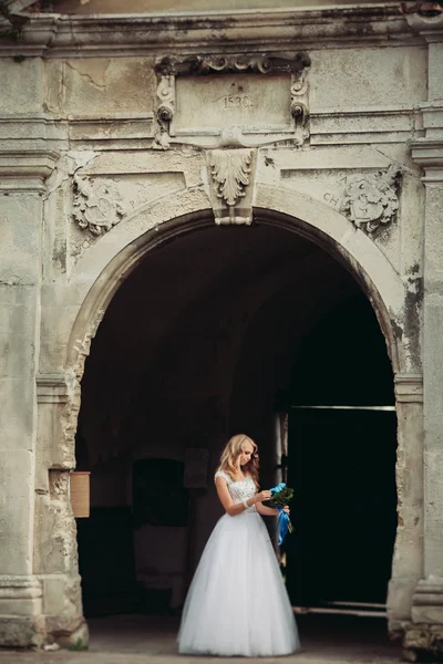 Όμορφη νεαρή ξανθιά νύφη με νυφική ανθοδέσμη που κάθονται στα σκαλοπάτια κάτω από πανέμορφες εγκαταστάσεις — Φωτογραφία Αρχείου