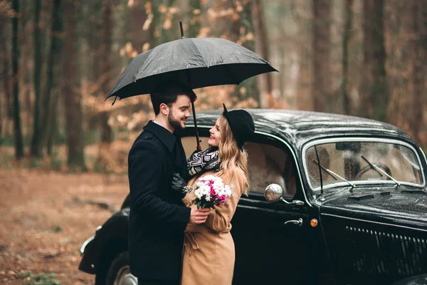 Casal amoroso elegante beijando e abraçando em uma floresta de pinheiros perto de carro retro — Fotografia de Stock