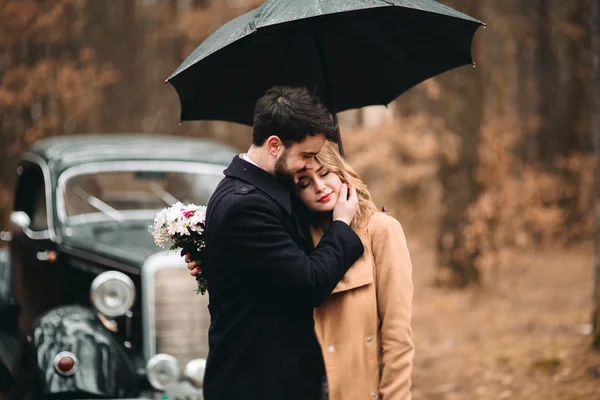 Casal amoroso elegante beijando e abraçando em uma floresta de pinheiros perto de carro retro — Fotografia de Stock