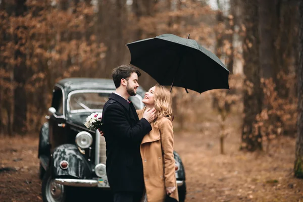 Casal amoroso elegante beijando e abraçando em uma floresta de pinheiros perto de carro retro — Fotografia de Stock