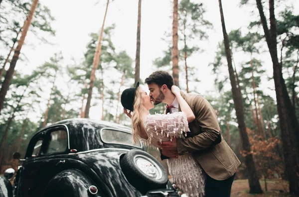 Şık Loving düğün çift öpüşme ve retro car yakınındaki bir çam ormanı içinde sarılma