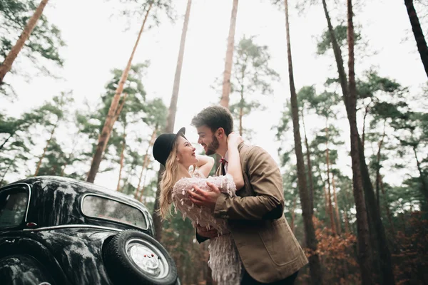 Стильна любляча весільна пара цілується і обіймається в сосновому лісі біля ретро-автомобіля — стокове фото