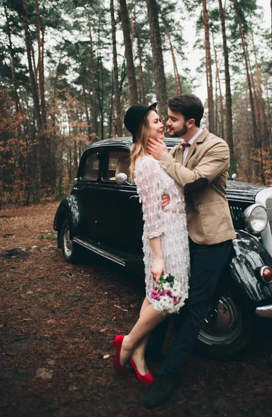 Elegante pareja de boda cariñosa besándose y abrazándose en un bosque de pinos cerca de coche retro —  Fotos de Stock