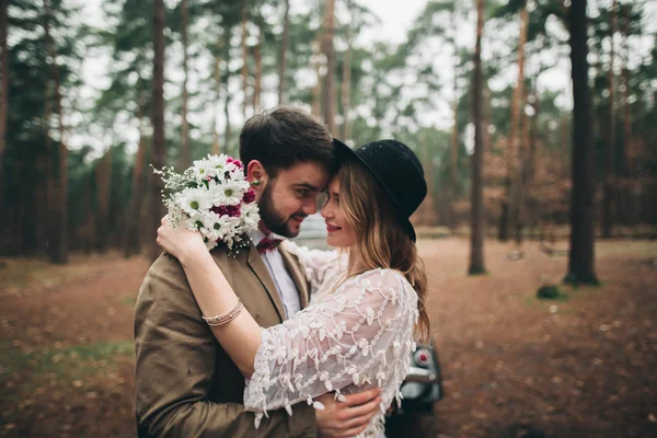 Stilvoll liebende Hochzeitspaar küsst und umarmt sich in einem Kiefernwald in der Nähe von Retro-Auto — Stockfoto