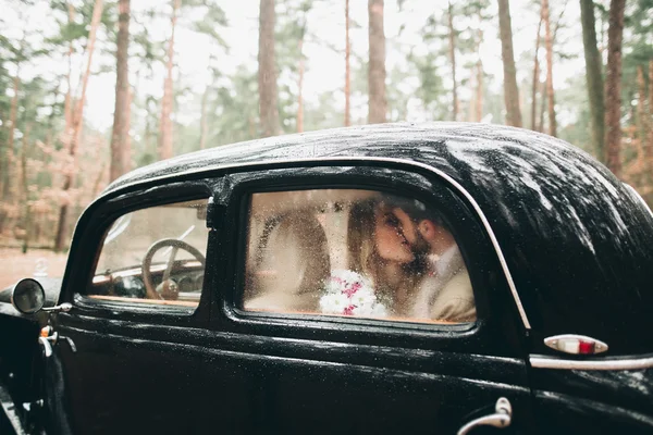 Casal amoroso elegante beijando e abraçando em uma floresta de pinheiros perto de carro retro — Fotografia de Stock