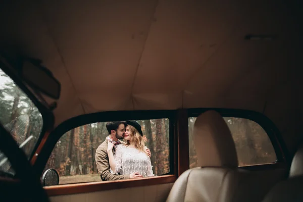 คู่แต่งงานที่มีสไตล์ จูบและกอดกันในป่าสนใกล้รถย้อนยุค — ภาพถ่ายสต็อก