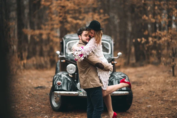 Casal amoroso elegante beijando e abraçando em uma floresta de pinheiros perto de carro retro — Fotografia de Stock