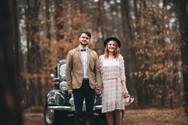 Şık Loving düğün çift öpüşme ve retro car yakınındaki bir çam ormanı içinde sarılma