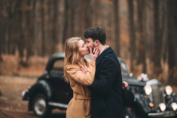 Casal amoroso elegante beijando e abraçando em uma floresta de pinheiros perto de carro retro — Fotografia de Stock